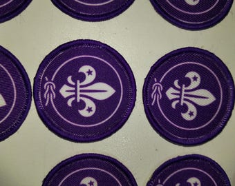 Może przedstawiać: Kolekcja okrągłych haftowanych naszywek z fioletowym tłem i białym emblematem lilii. Każda naszywka przedstawia centralny biały wzór lilii otoczony cienką białą okrągłą obwódką oraz detalem w postaci zawiązanego sznurka.