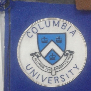 May include: A white and blue circular patch with the words "Columbia University" in a silver circle. The patch features a blue shield with a white chevron and three crowns.