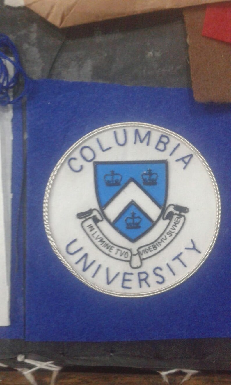 May include: A white and blue circular patch with the words "Columbia University" in a silver circle. The patch features a blue shield with a white chevron and three crowns.