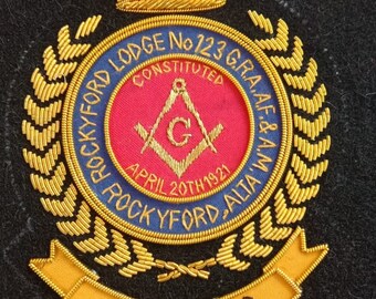 Może przedstawiać: Złota naszywka haftowana z niebieskim i złotym okręgiem. Okrąg zawiera tekst "Rockyford Lodge No 123 G.R.A.A.F. & A.W. Constituted April 20th 1921 Rockyford Alta'W" oraz czerwony kwadrat ze złotym kompasem i kątomierzem. Naszywka ma również złotą koronę i złotą wstęgę z tekstem "100 Years".