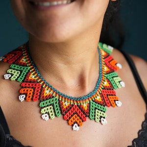 May include: A colourful beaded necklace with a geometric pattern in shades of green, orange, red, and white. The necklace is made of small beads and has a unique, handcrafted look.