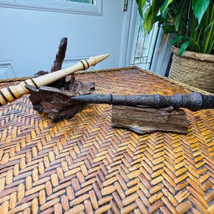 May include: Two handcrafted wooden wands rest on a decorative wooden base. One wand is light-colored with black bands, the other is dark brown with a textured surface. The wands are displayed on a woven table.