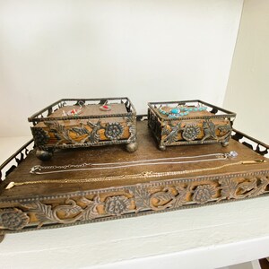 May include: A decorative metal tray with intricate floral designs and three legs. The tray has two smaller square trays with floral designs and legs. The smaller trays hold jewelry, including earrings, a necklace, and a bracelet.