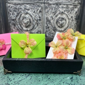 May include: A black wooden tray holds colorful envelopes and a card, all adorned with delicate floral embellishments. The envelopes are pink, green, and yellow, with matching floral accents. The card features multiple flowers.