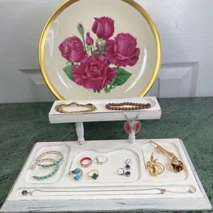 May include: White wooden jewelry organizer with two tiers. The top tier has two compartments for bracelets and the bottom tier has four compartments for rings, earrings, and a necklace. There are several pieces of jewelry in the compartments, including turquoise, silver, and gold.