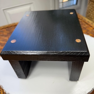 Könnte beinhalten: Ein kleiner, rechteckiger, schwarzer Holzschemel mit vier kleinen, runden, hellfarbigen Akzenten auf der Oberseite. Der Hocker hat ein schlichtes Design mit zwei stabilen Beinen auf jeder Seite.