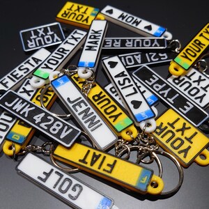 May include: A collection of colourful keychains with various names and words printed on them, including "FIESTA", "JWK 428V", "JENNIN", "YOUR TXT", "MARK", "LISA", "MOM", "521 A01", "YOUR RE", "E TYPE", "GOLF", and "FIAT".