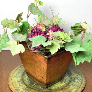 May include: A square copper-toned metal planter with a subtle polka dot pattern, filled with an arrangement of artificial green grape leaves, curling tendrils, and deep magenta-colored dried flowers that spill over the top edges.