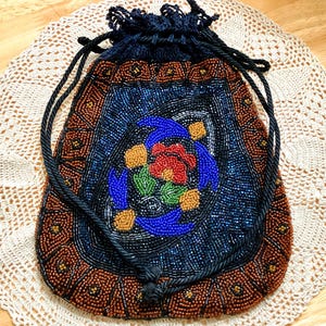 May include: A drawstring pouch featuring intricate beadwork in shades of brown, black, blue, red, green, and yellow. The bag displays a central floral motif surrounded by a geometric border, finished with a black cord closure at the top.