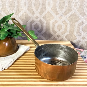 May include: A copper saucepan with a long brass handle. The interior of the pan shows signs of use. The pan is sitting on a wooden surface. A copper bowl and greenery are in the background.