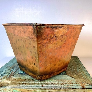 May include: A square-shaped copper planter with a tapered design and a weathered, oxidized patina. The exterior surface features a repeating pattern of faint, circular indentations across the metallic orange and brown body of the vessel.
