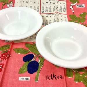 May include: Two white, oval-shaped bowls are displayed on a pink cloth with a calendar print and botanical illustrations. The cloth features text including "SLOE" and "ROWANBERRY".