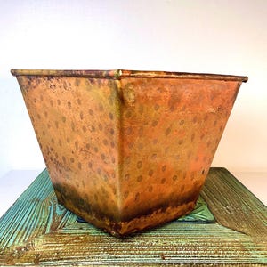 May include: A square-shaped copper planter featuring a tapered design and a weathered, mottled surface texture. The metal displays varied shades of orange, brown, and gold with a repeating pattern of faint, darker circular spots across the exterior.