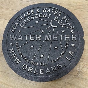Puede incluir: Una tapa de contador de agua redonda de hierro fundido gris oscuro. La tapa presenta letras en relieve que dicen "SEWERAGE & WATER BOARD CRESCENT BOX WATER METER NEW ORLEANS, LA." e incluye una luna creciente y estrellas.
