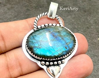 Puede incluir: Un colgante de plata con una piedra preciosa de labradorita ovalada con iridiscencia azul. La piedra está engastada en un bisel dentado con borde de cuentas, un asa de alambre trenzado y un pequeño acento de plata en forma de corazón. El texto KaviArty es visible.