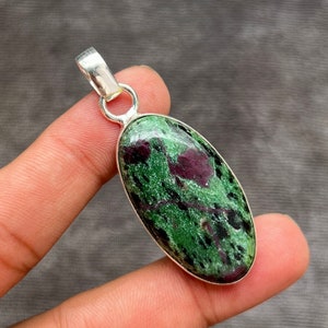 May include: A silver pendant featuring a green and red oval gemstone with a mottled pattern. The pendant is on a finger.
