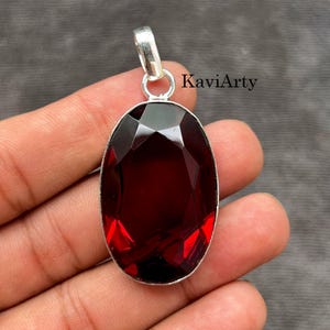 May include: An oval-shaped, deep red faceted gemstone pendant set in a silver-toned metal frame with a top bail. The text KaviArty is visible in the upper right corner. The pendant features a polished surface with geometric facets reflecting light.