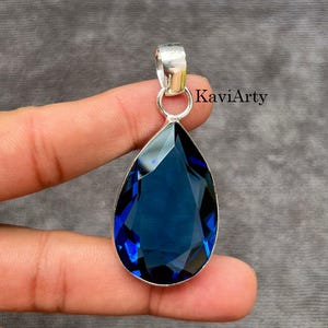 Peut inclure: Un pendentif en forme de goutte avec une pierre précieuse facettée bleu profond montée sur une bélière en métal argenté. Le pendentif porte l'inscription KaviArty en lettres noires. La pierre présente une surface polie à facettes.