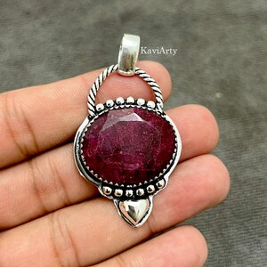 May include: A pendant featuring a large, oval-shaped, faceted deep red gemstone set in a silver-toned metal frame. The setting includes a twisted rope-like border, a series of small silver spheres, and a smooth heart-shaped accent at the base.