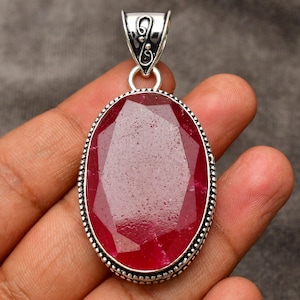 Pode incluir: Um pingente de prata com uma grande pedra de rubi facetada de forma oval. O pingente tem um design detalhado e ornamentado ao redor da borda da pedra.