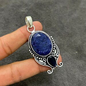 Op de afbeelding: Een zilveren hanger met een grote ovale blauwe saffier edelsteen en een kleinere peer-vormige blauwe saffier edelsteen. De hanger heeft een ingewikkeld zilveren ontwerp.