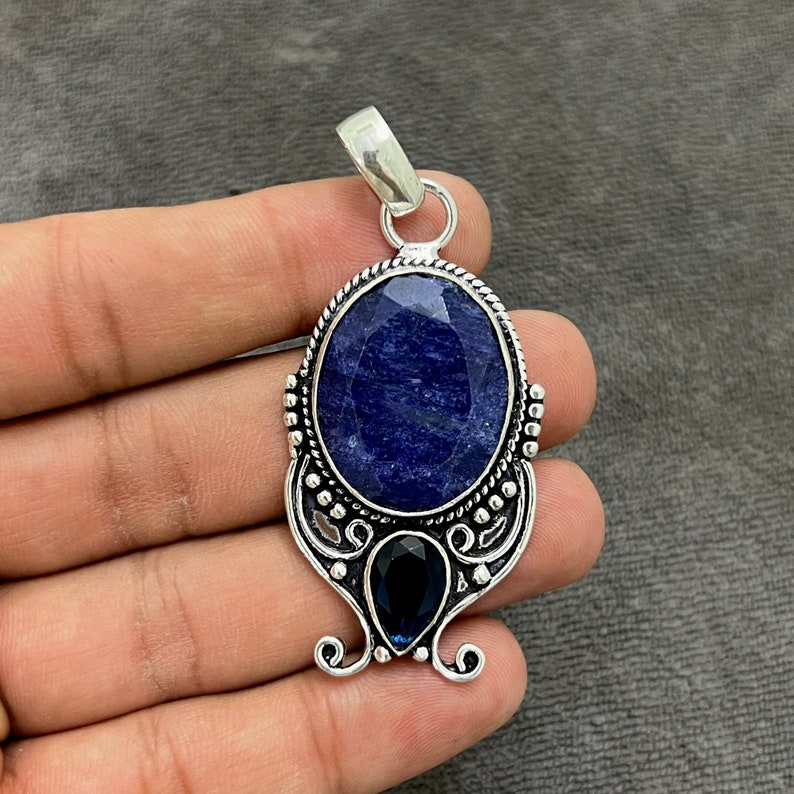 Op de afbeelding: Een zilveren hanger met een grote ovale blauwe edelsteen omgeven door een decoratief zilveren frame. Een kleinere blauwe edelsteen is onder de grotere geplaatst.