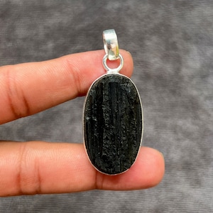 Pode incluir: Pingente de turmalina preta com uma fivela de prata. A pedra de forma oval tem uma textura estriada.