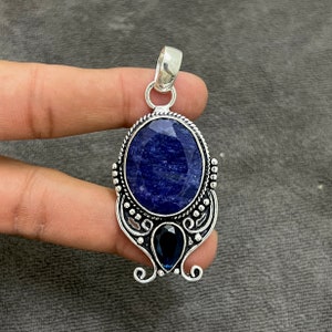 Op de afbeelding: Een zilveren hanger met een grote ovale blauwe edelsteen en een kleinere peer-vormige blauwe edelsteen. De hanger is versierd met ingewikkelde zilveren details.