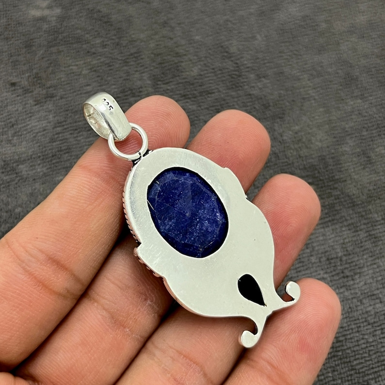 Op de afbeelding: Zilveren hanger met een blauwe edelsteen gezet in een gedetailleerd zilveren frame. De hanger heeft een lus om aan een ketting te bevestigen.