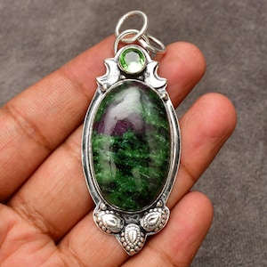 May include: A silver pendant with an oval-shaped, green and maroon stone. A smaller, round green stone sits above the oval stone. The pendant has a decorative silver design around the stones and a silver loop at the top.