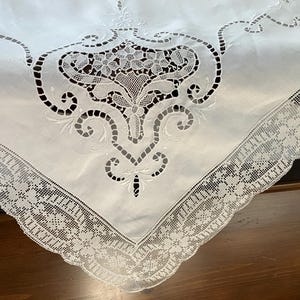 Pode incluir: Uma toalha de mesa branca com bordado recortado intrincado num motivo floral central e uma borda larga de renda festonada. O tecido exibe detalhes de costura minuciosos com secções abertas, criando um design decorativo no centro e bordas.