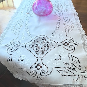 Puede incluir: Un camino de mesa rectangular blanco con intrincados bordados calados, motivos florales y un borde de encaje festoneado. Un jarrón redondo de cristal rosa translúcido con la parte superior ondulada y fruncida.
