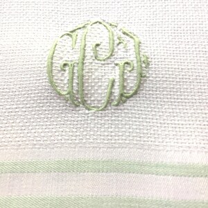 Op de afbeelding: Een witte handdoek van gestructureerde stof met een lichtgroen geborduurd monogram waarin de letters G, C en L in een cirkelvormig ontwerp zijn verweven. Het onderste deel van de handdoek toont twee horizontale lichtgroene strepen.