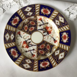 Può includere: Un piattino in porcellana blu e oro con un motivo floreale. Il piattino ha un centro bianco con un disegno floreale rosso. Il bordo del piattino è decorato con un motivo geometrico oro e blu.