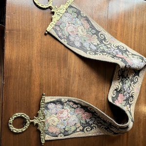 May include: Two decorative tapestry wall hangings with floral patterns and ornate brass ring holders. The tapestry features a black background with pink, blue, and cream-coloured flowers. The brass hardware adds a vintage touch.