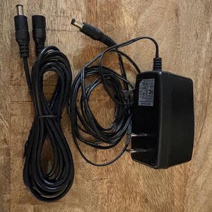 May include: A black power adapter with a two-prong plug and a DC power cord. The image shows a coiled black cable and two additional black cables with DC connectors. The items are on a wooden surface.