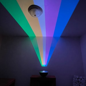 May include: A rainbow projector casts colorful light beams onto a white ceiling. The projector is a small, round device with a white base and a silver top. It is sitting on a table in a dark room.