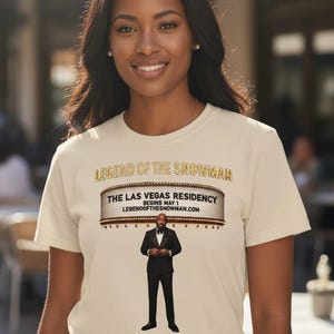 May include: A beige t-shirt featuring the text "LEGEND OF THE SNOWMAN" above a marquee design that reads "THE LAS VEGAS RESIDENCY BEGINS MAY 1 LEGENDOFTHESNOWMAN.COM". Below the marquee is an illustration of a man in a suit.
