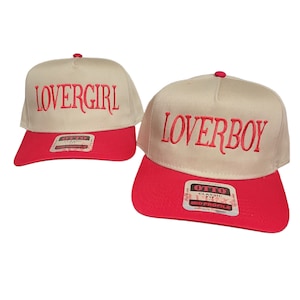 Puede incluir: Dos gorras de béisbol beige con viseras rojas. La parte delantera de las gorras tiene las palabras "Lovergirl" y "Loverboy" bordadas en rojo.