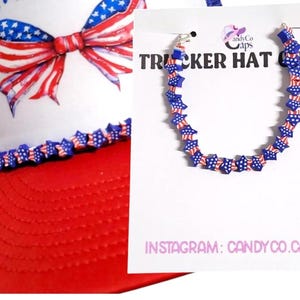 May include: A red and white trucker hat with a patriotic bow and star-shaped chain. The hat features a red brim and a white front panel with a bow design in red, white, and blue. A star-shaped chain is also visible.