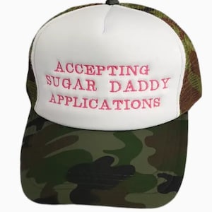 May include: A white trucker hat with a camouflage brim. The front of the hat has pink embroidered text that reads "ACCEPTING SUGAR DADDY APPLICATIONS".