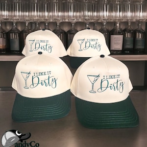 Puede incluir: Cuatro gorras de béisbol blancas con viseras verdes. Cada gorra tiene un gráfico de copa de martini y el texto "I like it Dirty".