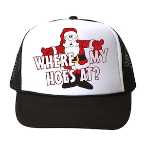 以下が含まれることがあります： 黒のメッシュバックが付いた白いトラッカーハット。ハットの前面には、サンタクロースの漫画の画像と「Where My Hoes At?」というテキストが描かれています