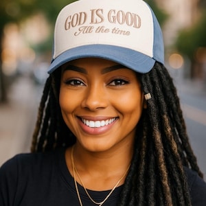 以下が含まれることがあります： 「GOD IS GOOD All the time」の刺繍が入った青とベージュのトラッカーハットを人が被っています。その人は笑顔で、黒いシャツと金のネックレスを身につけています。背景はぼやけています。