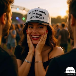 Puede incluir: Una gorra de béisbol blanca y negra con el texto "PRETTY GOOD AT BAD DECISIONS" es usada por una persona sonriente. La persona está rodeada de otras personas en un entorno al aire libre. La gorra tiene una visera negra y el texto es negro.