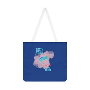 Op de afbeelding: Een blauwe tote bag met een witte band heeft een aquarelachtige illustratie van een zeeschildpad die zwemt met de tekst "Brave Little Turtle Love Out There".