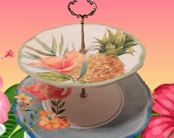 Puede incluir: Un soporte de servicio de dos niveles con un diseño de piña, flor de hibisco y flamenco. El soporte es blanco con hojas verdes y detalles azules. El nivel superior tiene un asa de metal plateado.