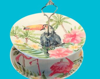 Puede incluir: Un soporte de servicio de dos niveles con un tema tropical. El nivel superior presenta un tucán con un pico naranja brillante y una flor de hibisco rosa. El nivel inferior presenta flamencos rosas y palmeras. El soporte tiene un asa plateada.
