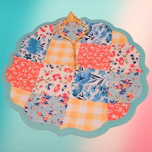 May include: A blue and white ceramic serving tray with a gold metal handle. The tray is decorated with a patchwork design of floral and checkered patterns.