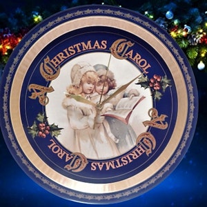 Puede incluir: Un reloj de Navidad azul y dorado con una imagen de dos niños cantando un villancico. La esfera del reloj dice "A Christmas Carol".